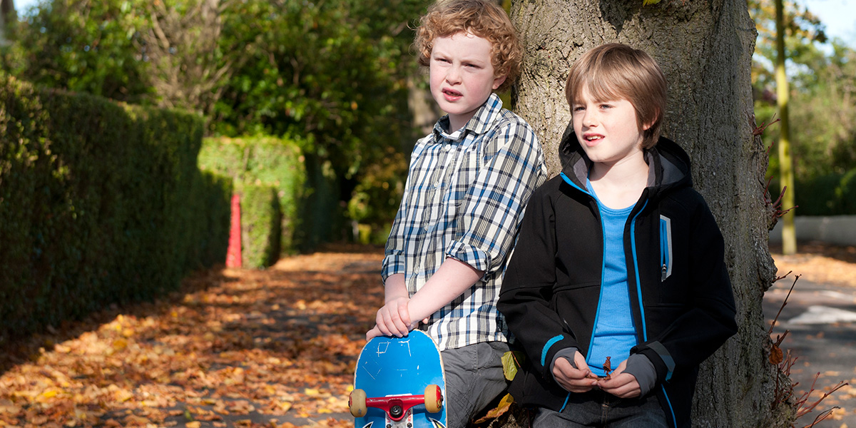 two boys with a skateboard standing beside a tree