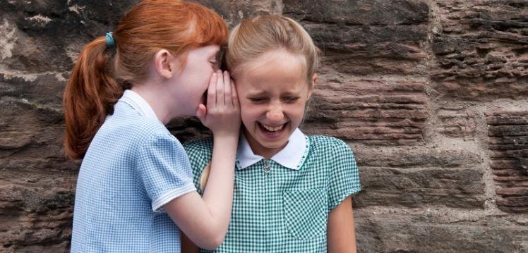 photograph of two primary school aged girls in different school uniforms whispering.