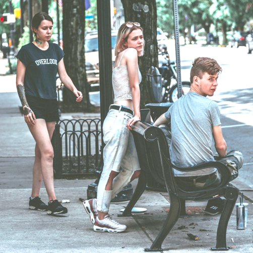 Two female teenagers are standing near a bench where a male is seated