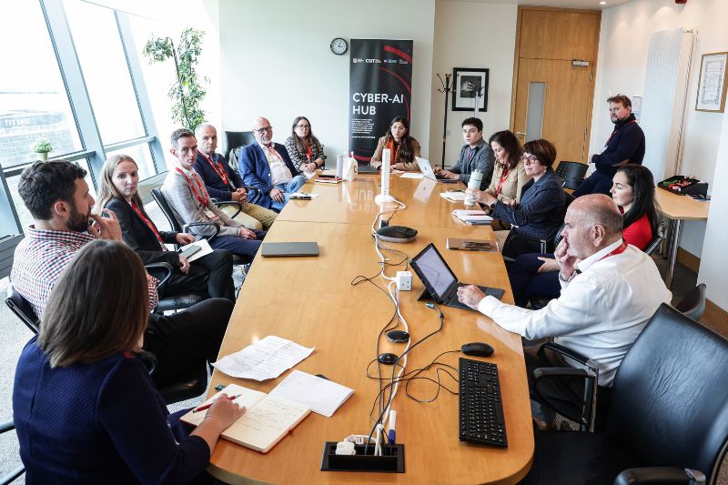 Group of researchers and industry visitors in discussion around a conference table at the CSIT, Queen’s University Belfast.