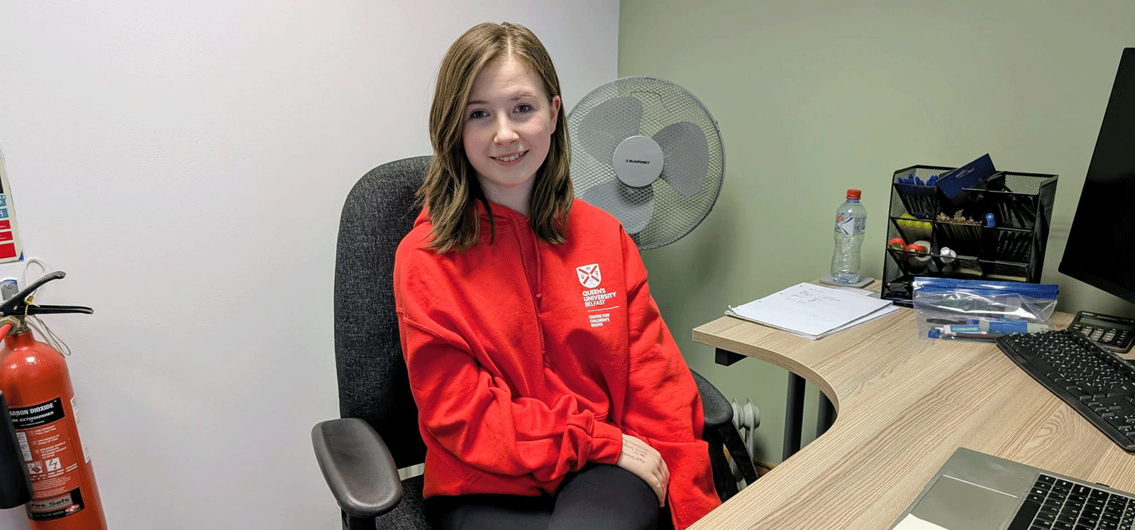 Sarah wearing a red Queen’s University Belfast hoodie, seated at a desk in an office and smiling at the camera.