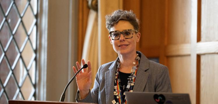 smiling woman with short hair and pattern-rimmed glasses, a grey jacket and chunky necklace, standing at lectern with wood panelling in the background