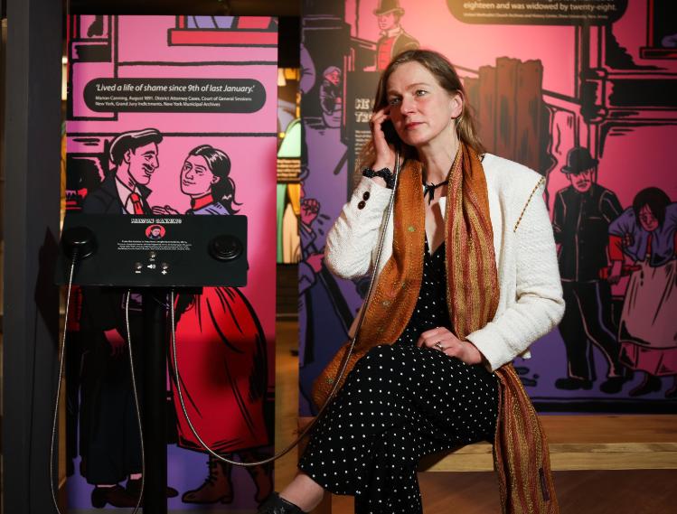 Woman wearing a white jacket, black and white polka dot dress and orange stripe scarf, sitting in a sound booth listening on an ear piece..