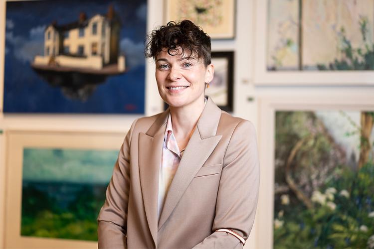 head and shoulders of Gail McConnell smiling and standing in front of a display of paintings