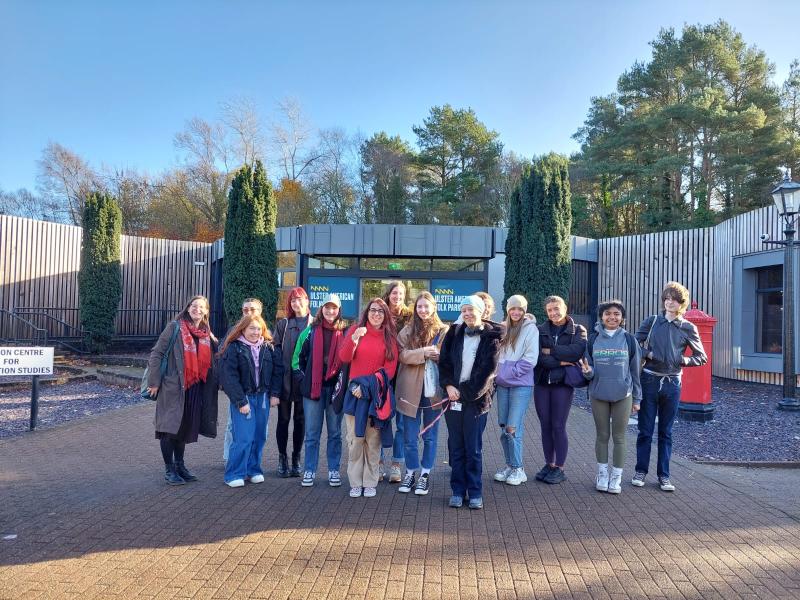 Sophie Cooper standing on the left of a group of smiling students in front of the Ulster American Folk Park museum building on a sunny day