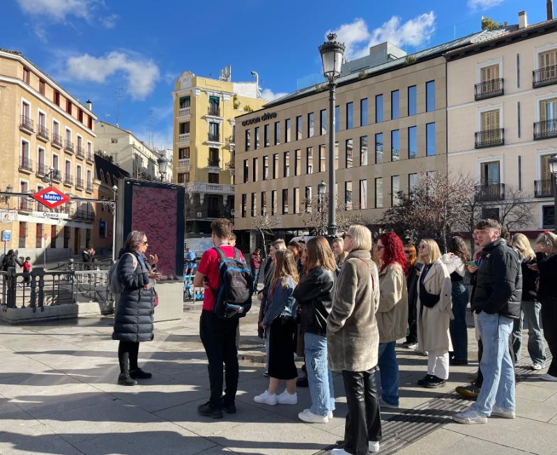 people standing in a small square outside a Madrid metro station listening to a tour guide. It is a bright day with sunshine.right