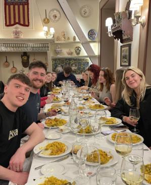 A group of smiling students seated at a long table ina restaurant with dishes of paella in front of them