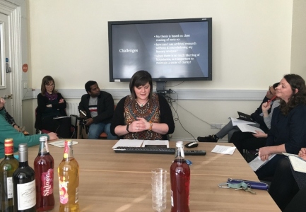 Seminar speaker delivering talk at a boardroom table with large display screen behind. Guests seated at same table with refreshments in the centre,