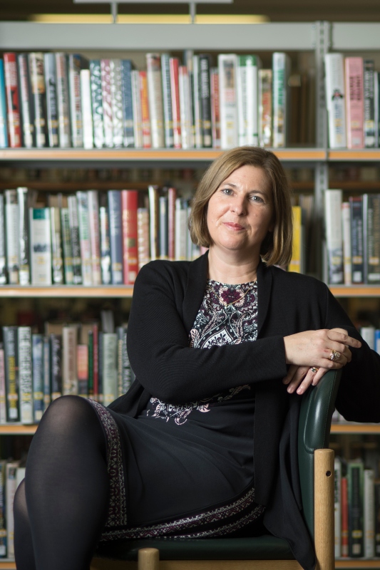 Janice Carruthers seated in front of a bookcase with left arm resting on arm of chair and right hand on top of left hand.