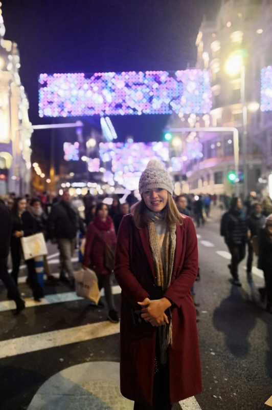 Erin McCombe wearing a red coat warm grey hat and matching scarf.  She's standing smiling in a busy shopping street illuminated with Christmas lights 