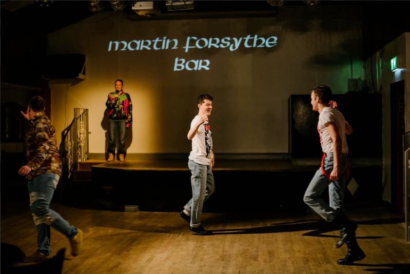 actors on stage, two happily greeting each other in the foreground, they are wearing jeans and white t-shirts. On the left one actor, wearing camoflage is exiting the picture and towards the back of the stage an actor wearing a brightly coloured top is singing underneath an illuminated sign saying Martin Forsythe Bar