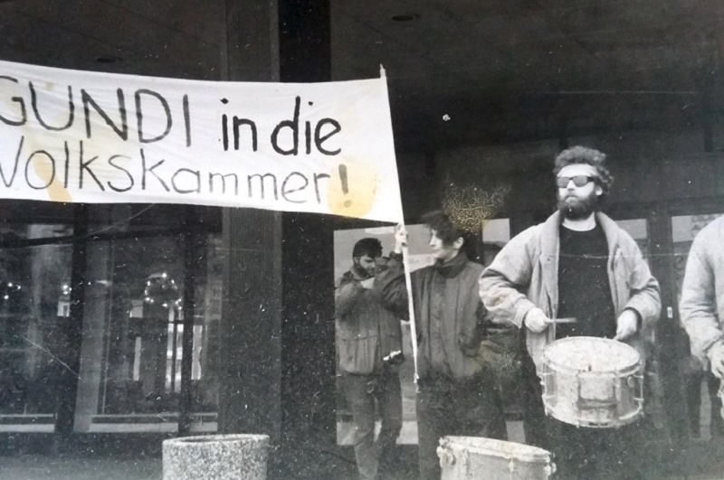 Black and white image of a protest.  Bearded man in foreground beating a drum, others behind holding banner reading 'GUNDI in die Volkskammer!'