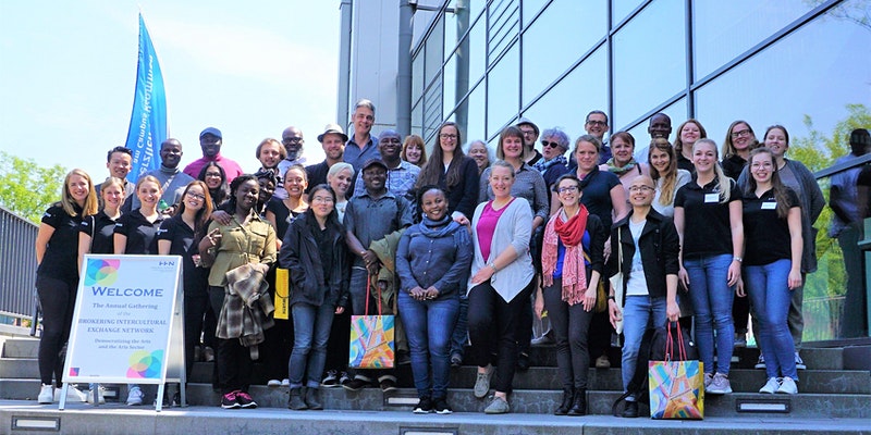 A group of smiling people outside in the sunshine standing on steps in front of a glass building. Sandwich board in the foreground reads Welcome to the Annual Gathering of the Brokering Intercultural Exchange Network.