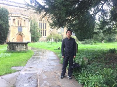 PhD Student profile photo. A male student standing outside in the lawn with an old building in the background, he is holding an umbrella