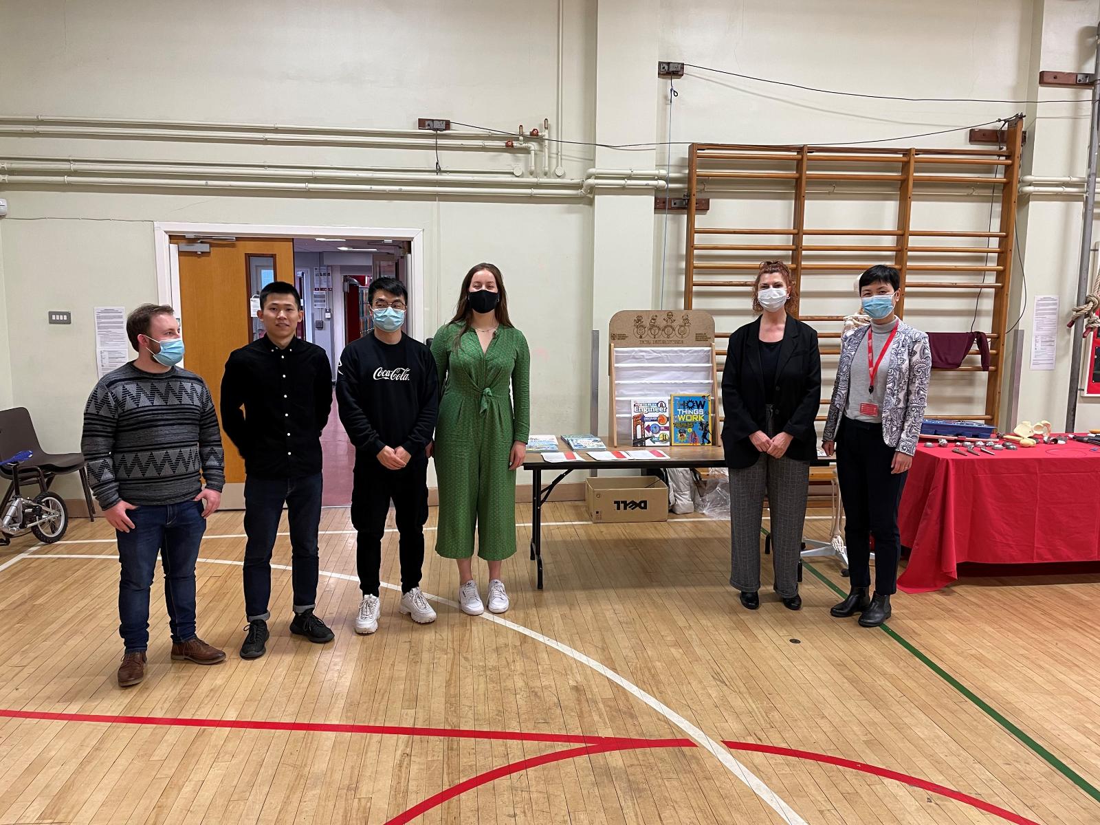 4 students and 2 staff members standing in a school sports hall in front of a table with STEM books on it