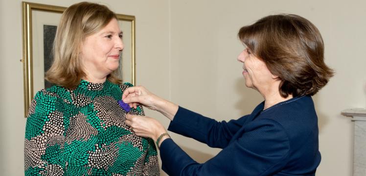 On thh left hand side, Janice Carruthers, in a green and blue dress, stands smiling while having the Chevalier dans l’Ordre des Palmes Académiques pinned to her dress by the French Ambassador to the UK, Her Excellency Mme Catherine Colonna.