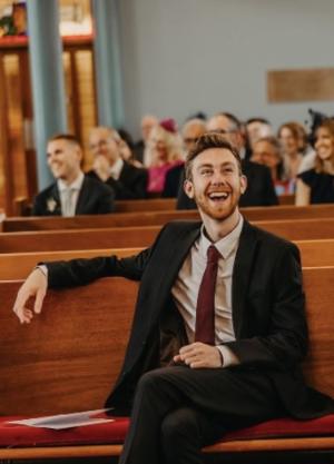 A male student sitting in a church bench looking very happy
