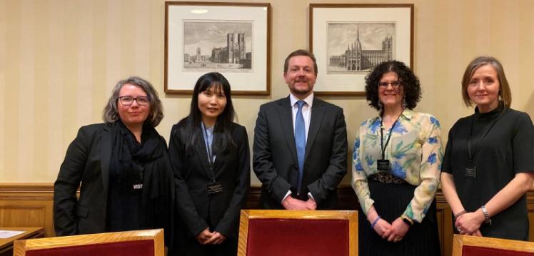 group of four females and one male in the centre, in a row against a stripey wall below two framed drawings of the Houses of Parliament