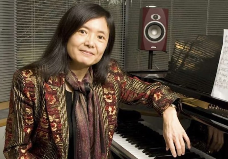 Noriko Manabe sitting in front of a piano, elbow resting on lid of instrument with sheet music behind