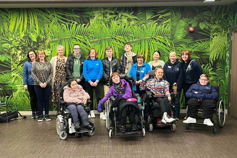 Back row of eleven standing young people and adults and front row of four young people and adults in wheelchars, with a jungle print wall behind the group