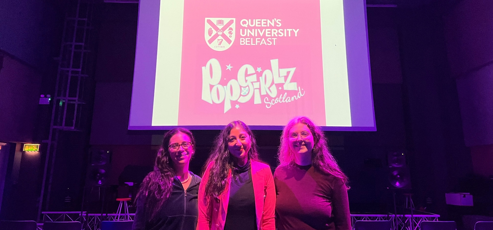 Three women standing in front of a large screen with logo on it