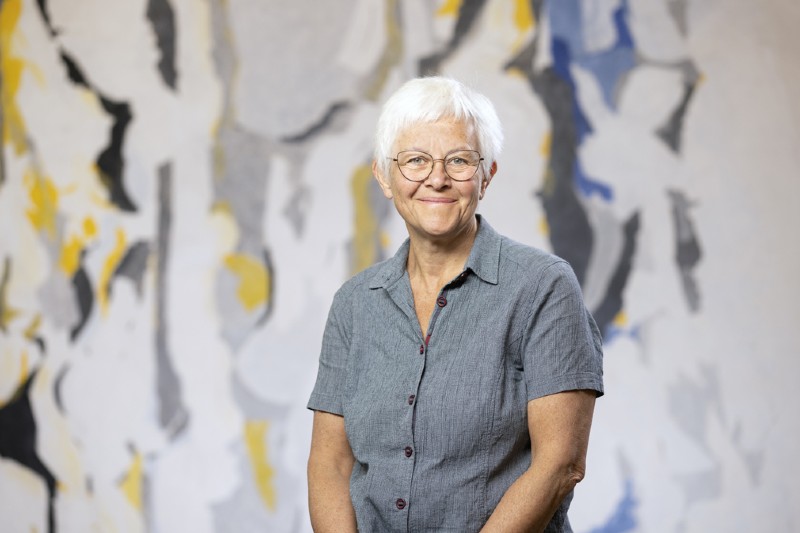 smiling woman with short grey hair and glasses, against a multi-coloured, abstract background wall