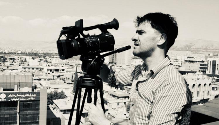Black and white image of Mark Cousins, wearing a striped shirt and filming using a video camera on a tripod. There is a cityscape in the background.