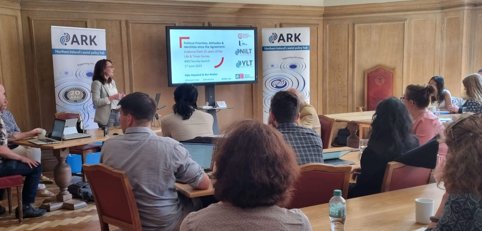 Katy Hayward presenting beside a screen with survey information on it, a wood panelled wall behind her and an ARK pop-up banner on either side of the photo