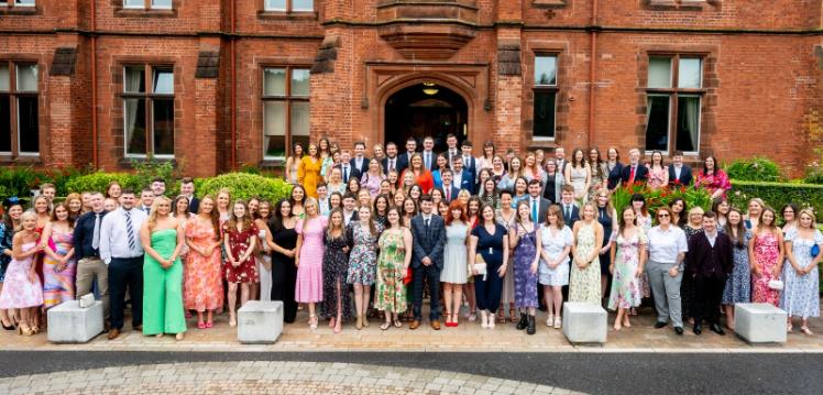 100+ smartly dressed men and women in their early twenties lined up in several rows on the steps leading down from the entrance to a large, red-brick building