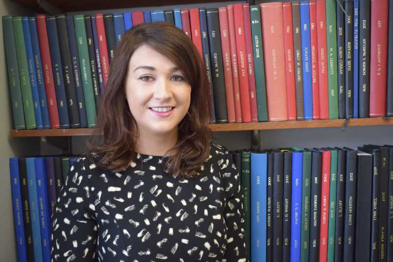 smiling young woman with shoulder length dark hair and a black spotty dress, with bookshelves behind her