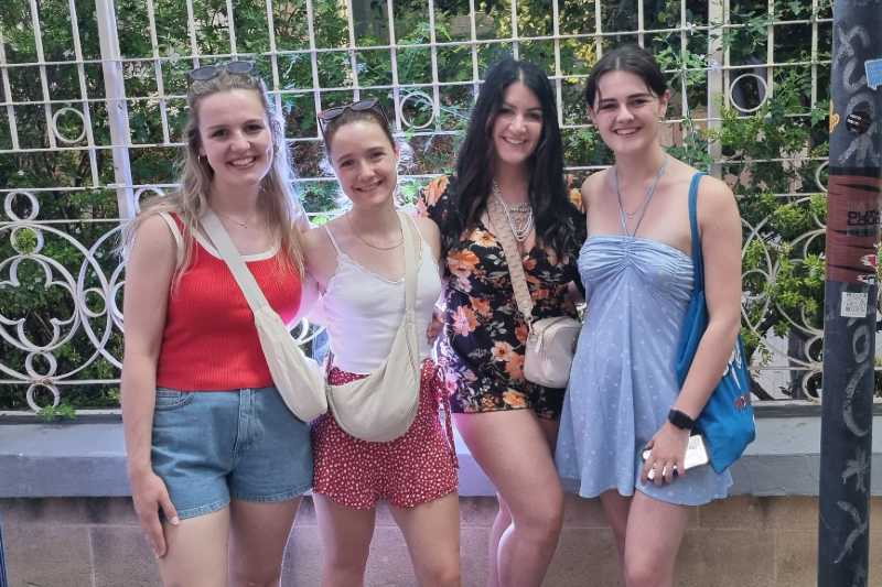 four smiling young women in summer clothes, against a low wall and railings