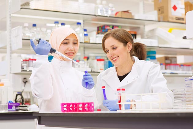 two female students in labcoats using a pipette dropper to add a substance to a test tube