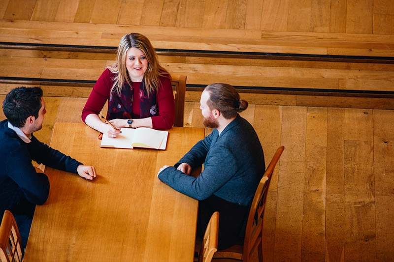 high angle view of three students sitting at a desk having a conversation with one of them taking notes