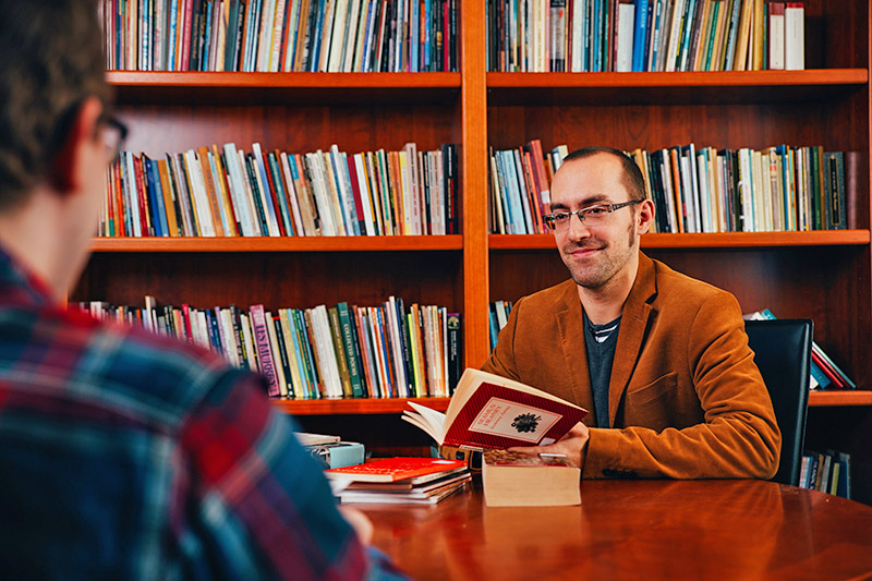 male student wearing glasses holding a book and having a conversation with another student sitting across from him at a desk