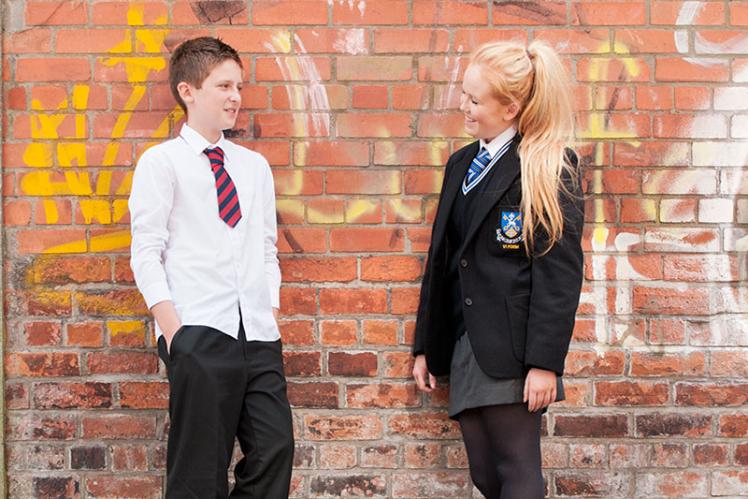 two post-primary pupils from different schools wearing different school uniforms standing against a brick wall covered in graffitti