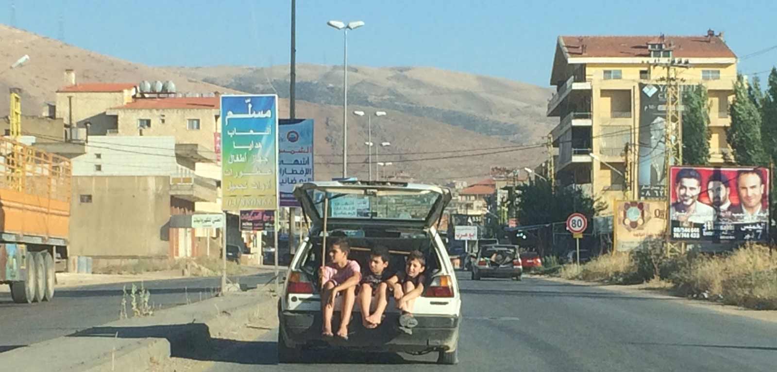 Three children sitting in the open boot of a car in a middle eastern town