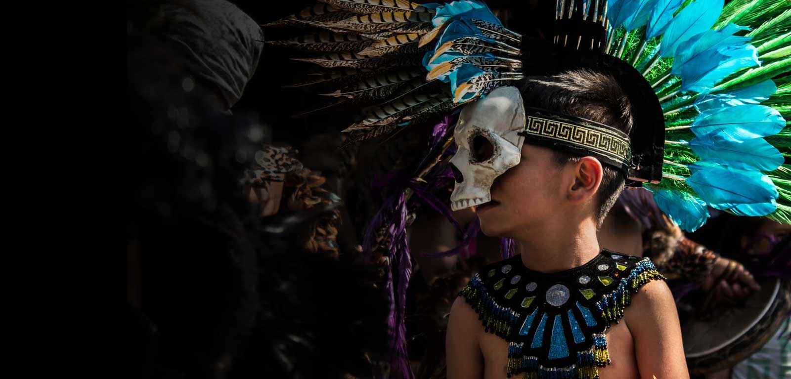 Picture of a young boy wearing a skull mask and colourful feathered headwear 