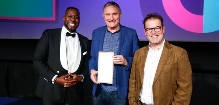 three smiling men standing in a row, with the central man holding an award