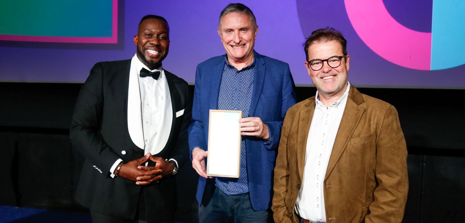 three smiling men standing in a row, with the central man holding an award