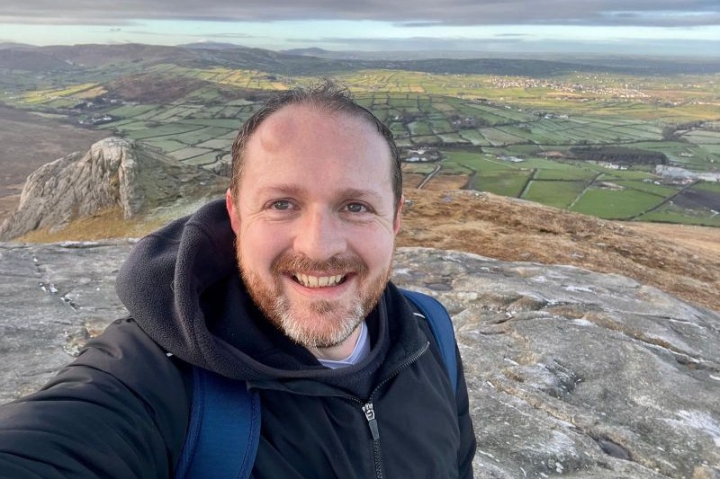 close up of smiling man with moustache and beard, with green hills behind
