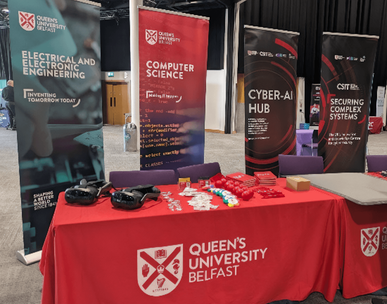 Exhibition booth setup for Queen’s University Belfast at an event. Four vertical banners display themes from the School of Electronics, Electrical Engineering and Computer Science: 'Electrical and Electronic Engineering', 'Computer Science', 'Cyber-AI Hub', and 'Securing Complex Systems'. A red tablecloth with the Queen’s University Belfast logo covers the display table.