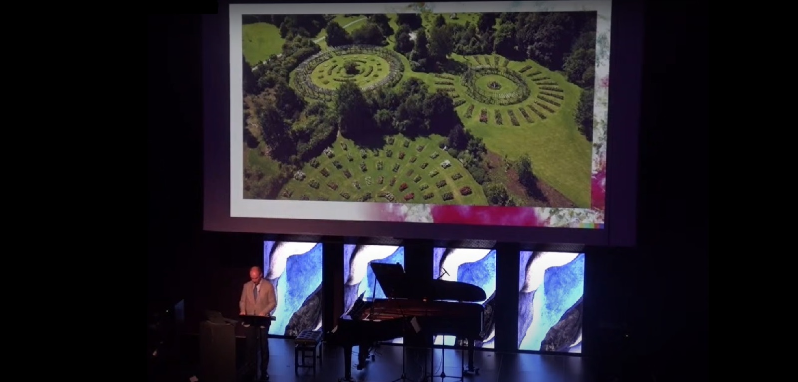 Man standing on a stage delivering a lecture in front of a large screen