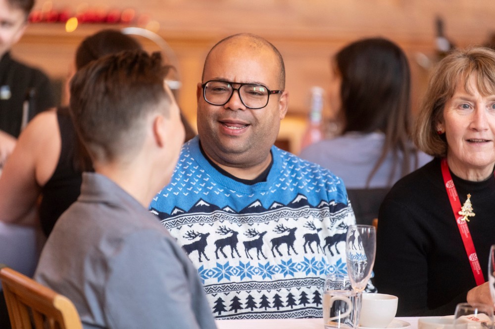 swarthy man in Christmas jumper chats to colleagues at a table at a seated indoor event