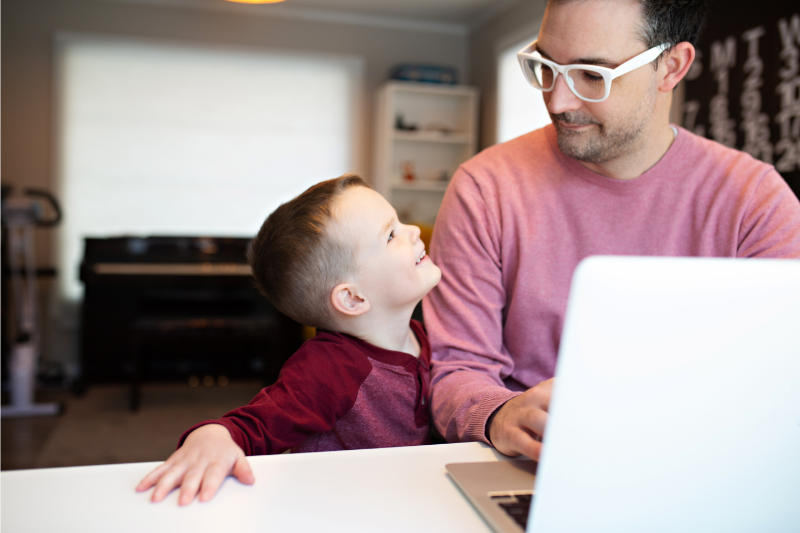 dad working on laptop with young son smiling up at him