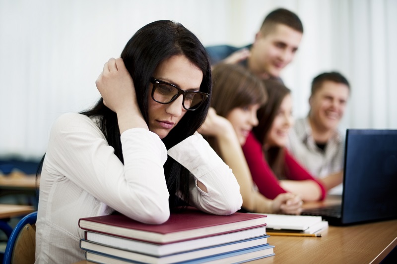 Tense-looking dark-haired young woman with glasses sitting at a desk leaning on a stack of books and covering her ears, beside a group of happy young people in the background looking at a laptop