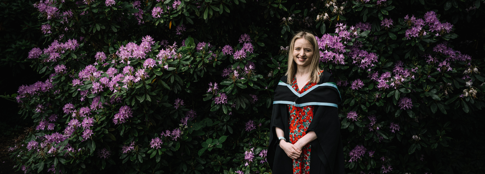 female student in graduation gown standing near some flowering plant outside