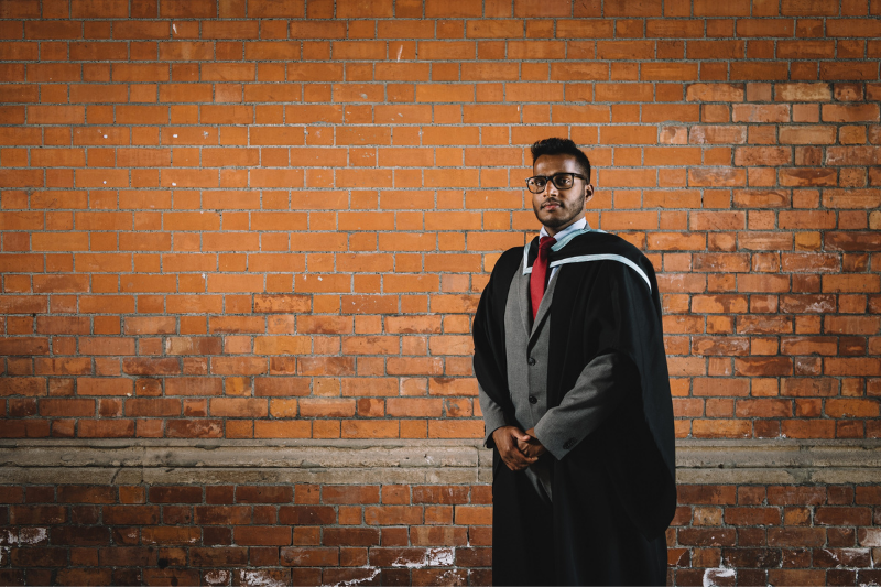 male graduate in graduation gown backdrop is a brick wall
