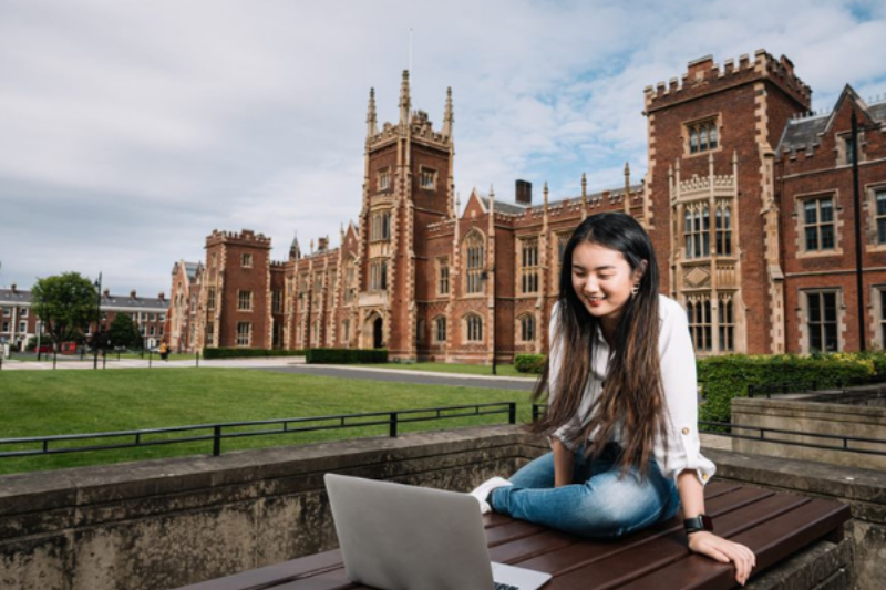 female graduate sitting outside Queen's University on a laptop