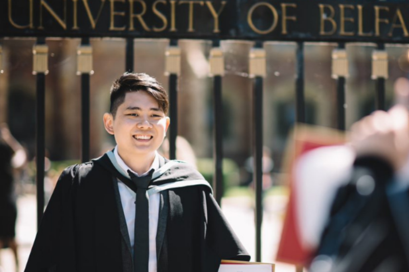 male student on his graduation day standing at gate of Queen's University with degree certificate