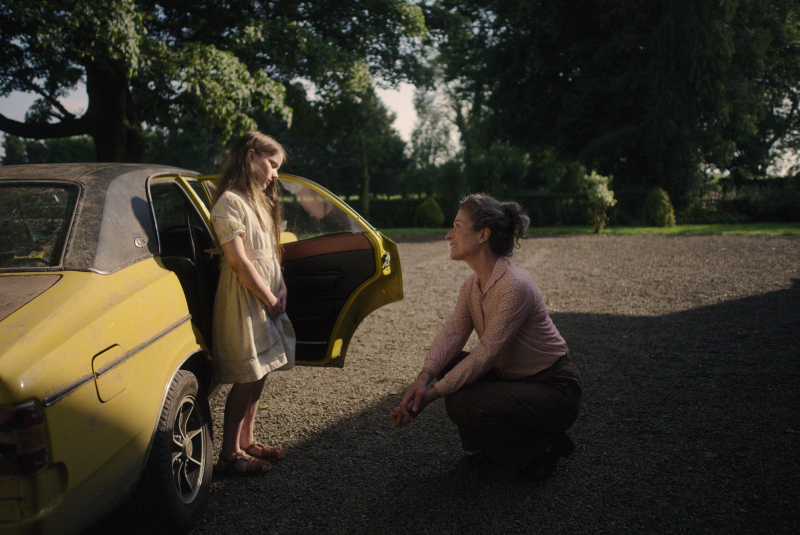 still from the film The Quiet Girl - a young girl in a pale yellow/cream dress standing outside an opened car door and talking to an older woman who is sitting on her haunches on the ground